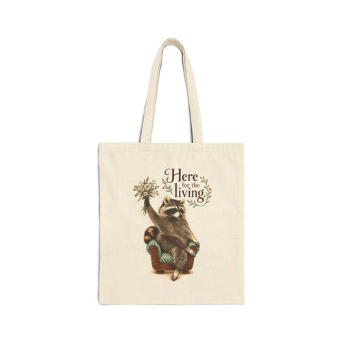 Natural canvas tote with Taco the raccoon beneath “Here for the living” lettering and a woodland flower bouquet.