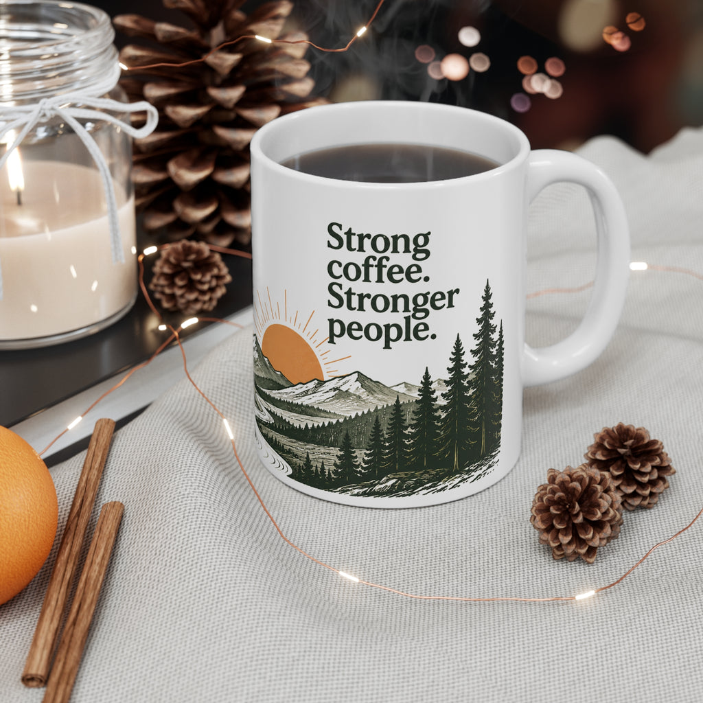 White ceramic “Strong coffee. Stronger people.” mug with mountain and evergreen sunrise artwork, styled with pinecones, a candle, and cinnamon sticks on a neutral table.